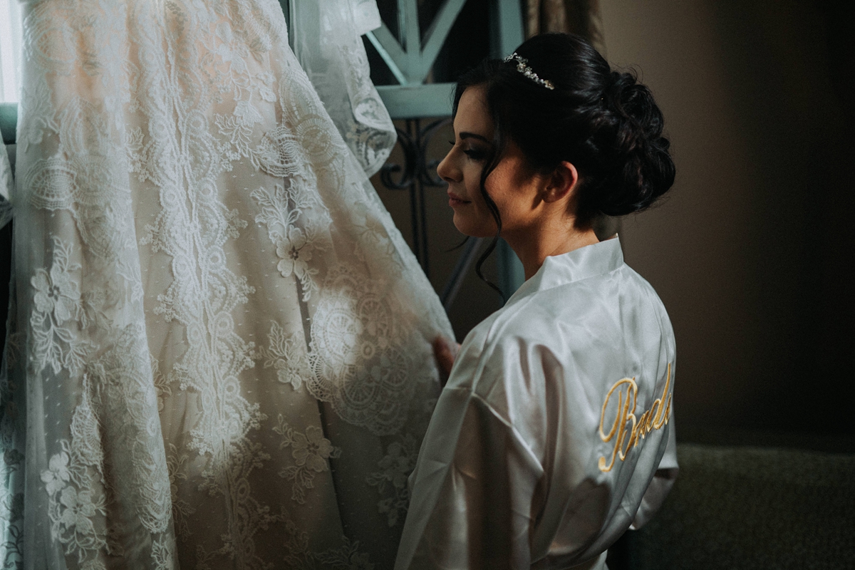 Bride in white silk robe smiling at hanging wedding dress