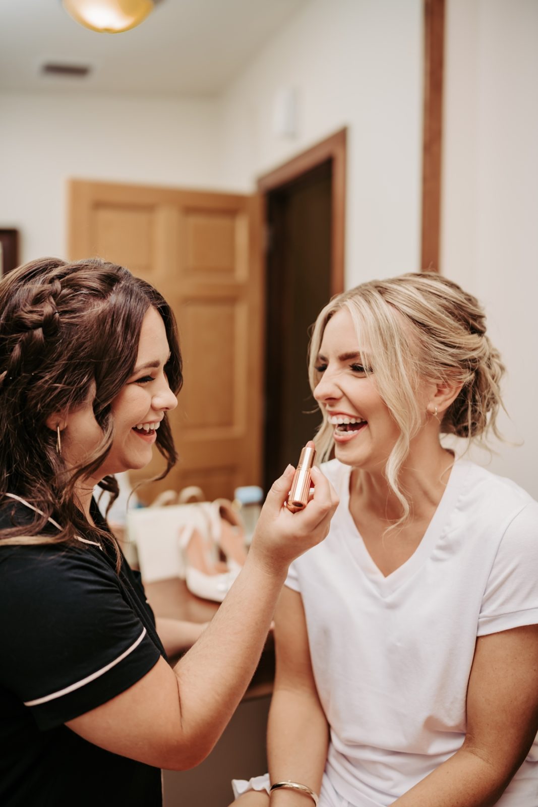 Bridesmaid helping bride apply lipstick and laughing