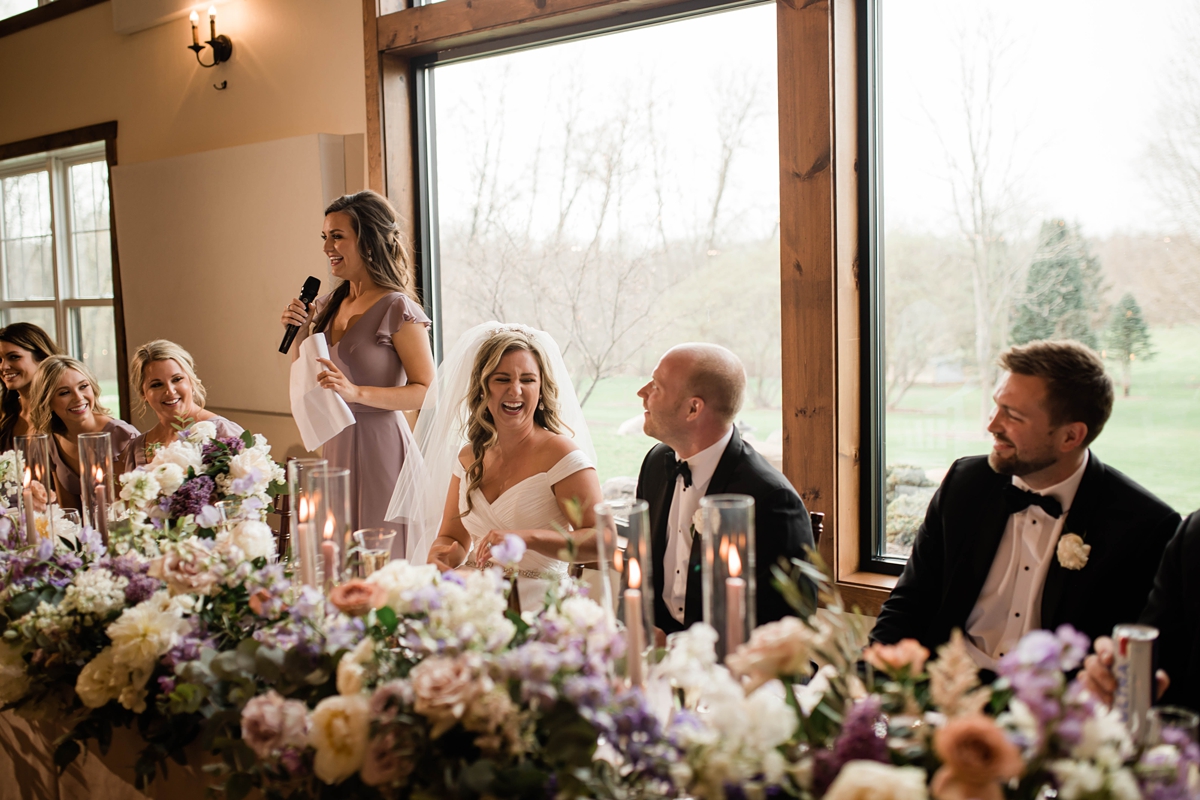 Bridesmaid giving speech at reception while bride laughs.