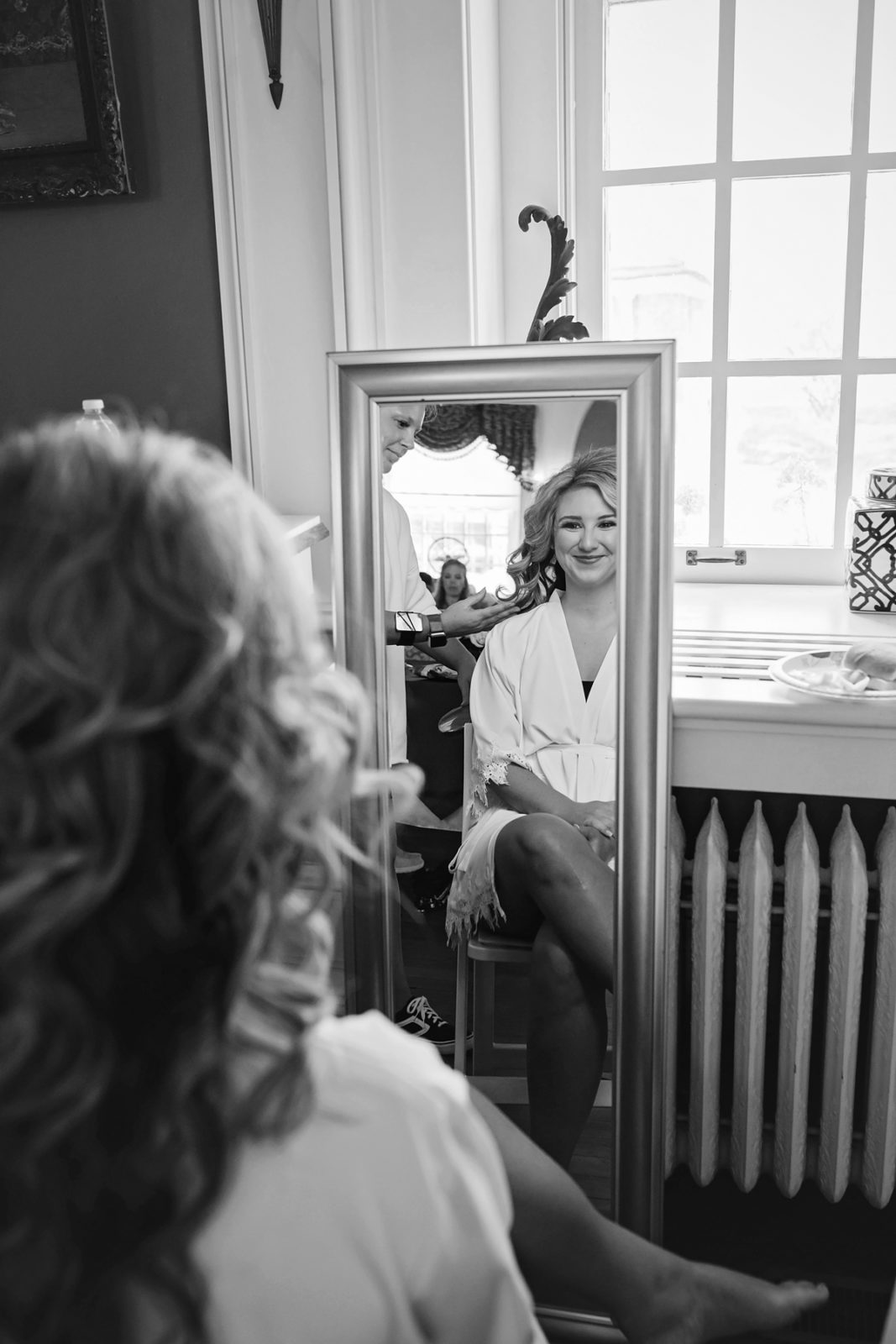 Black and white photo of bride getting ready while smiling at herself in mirror