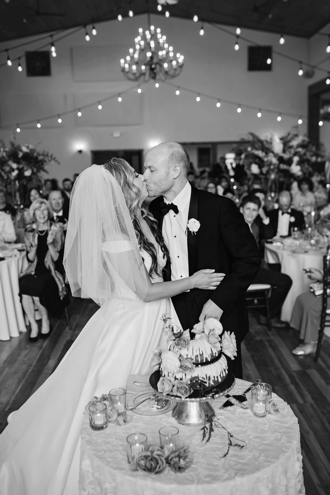 Black and white image of bride and groom kissing while cutting wedding cake.